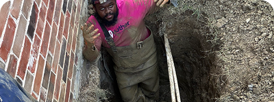 Man wearing waders stands inside a deep rectangular hole next to a brick wall, holding two white pipes, appearing to explain or gesture.