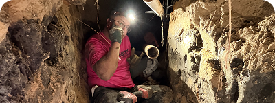 A person wearing a headlamp and a pink shirt working inside a narrow underground tunnel, holding a pipe and surrounded by dirt walls.