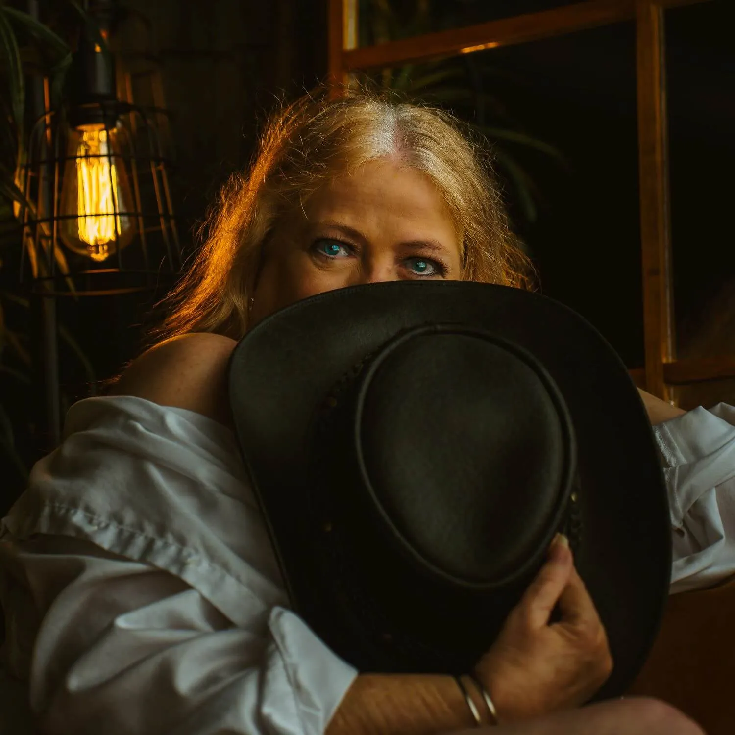 Senior boudoir set, in unbuttoned light blue shirt, playfully hiding face with black cowgirl hat.