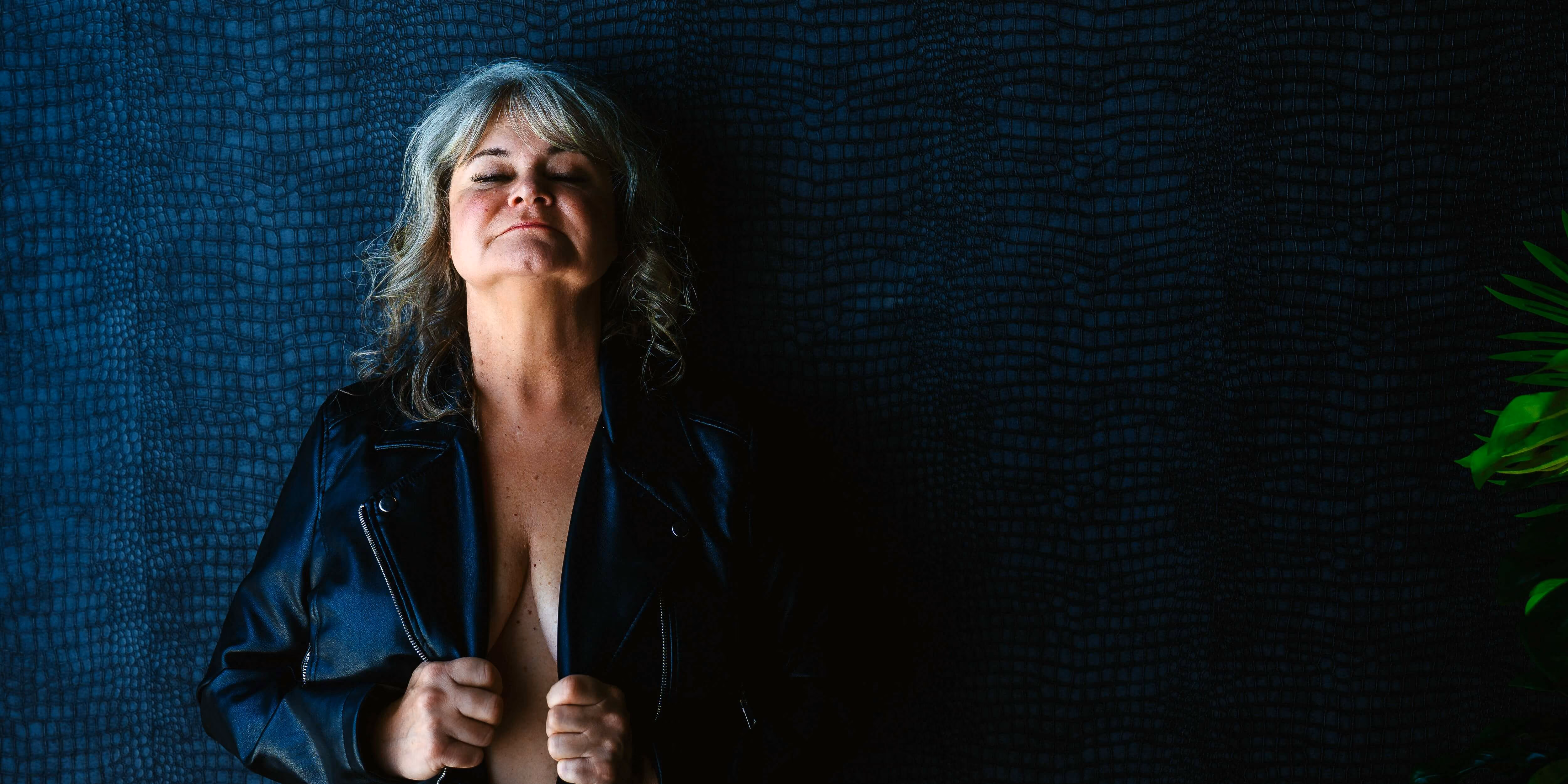 Senior woman in an open black leather jacket against a blue patterned wall.