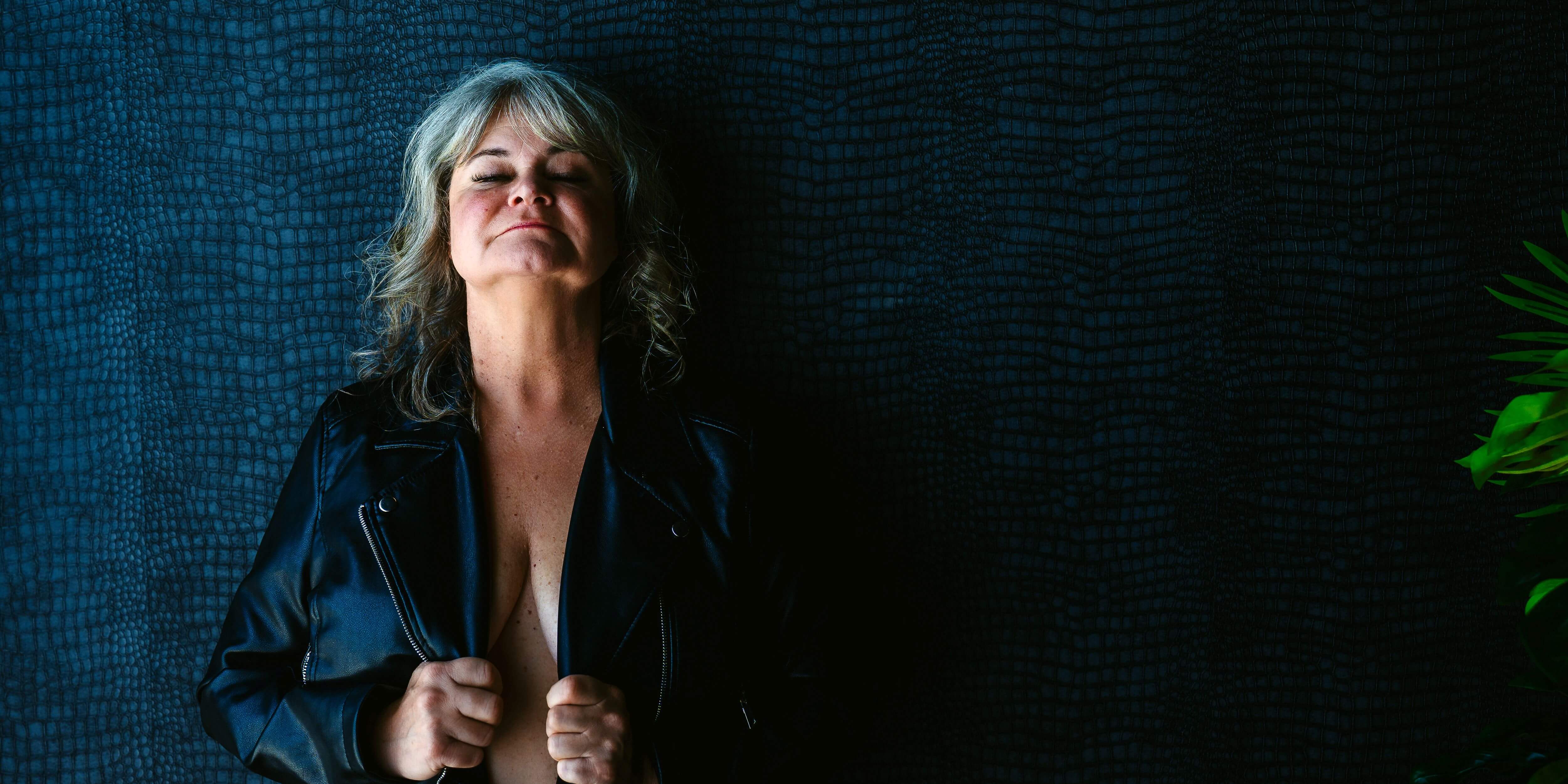 Confident senior woman posing shirtless, in black leather jacket, against a blue patterned wall. 