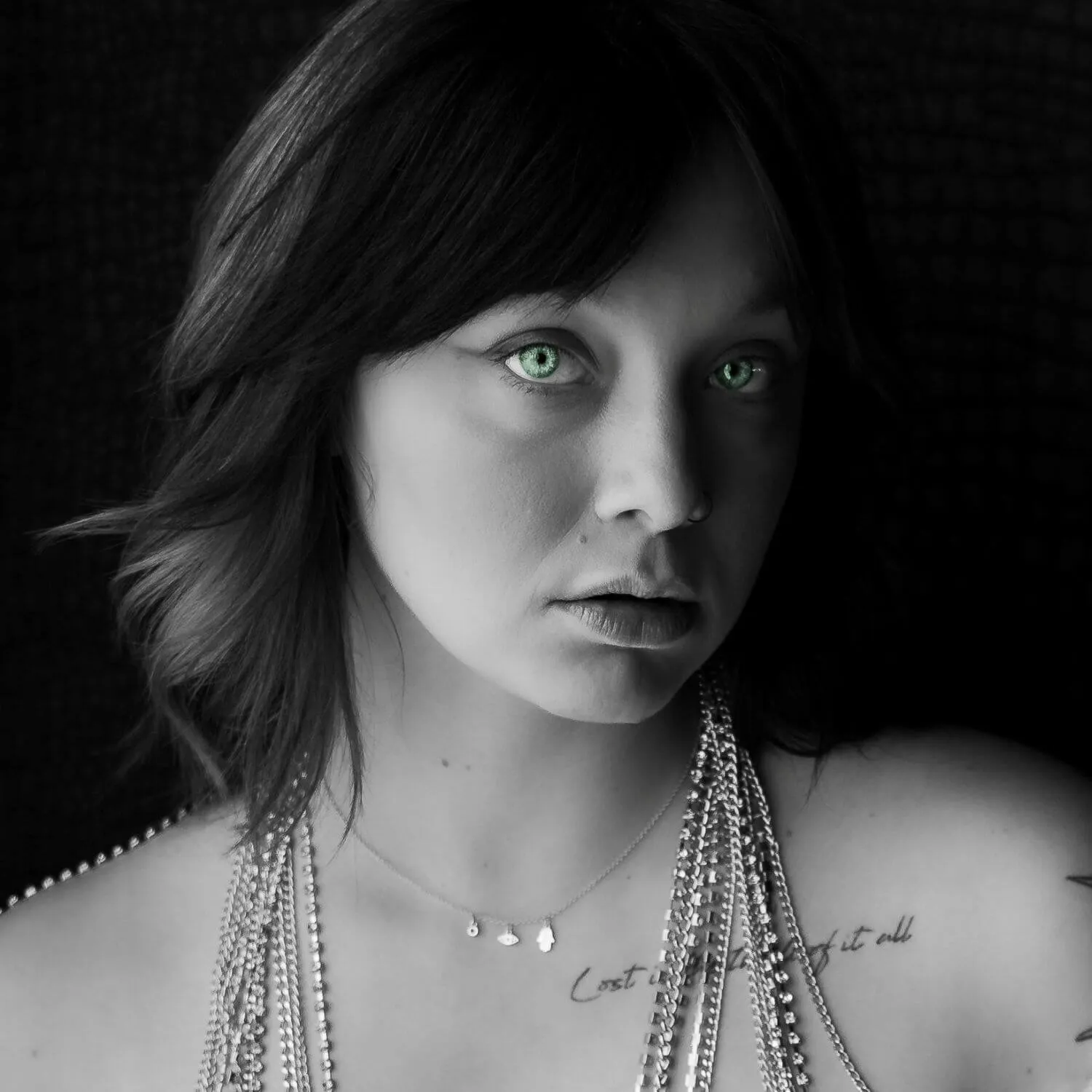 Woman staring into camera, adorned with multiple necklaces. Black and white, with striking green eyes.