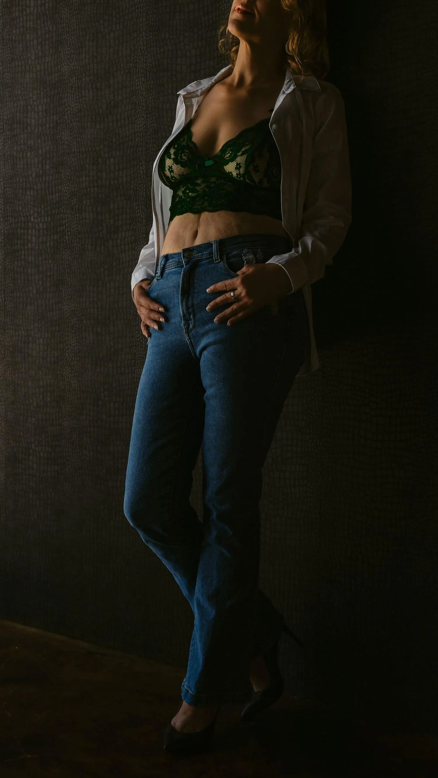 Confident, mature woman leaning against wall in jeans and green lingerie top.