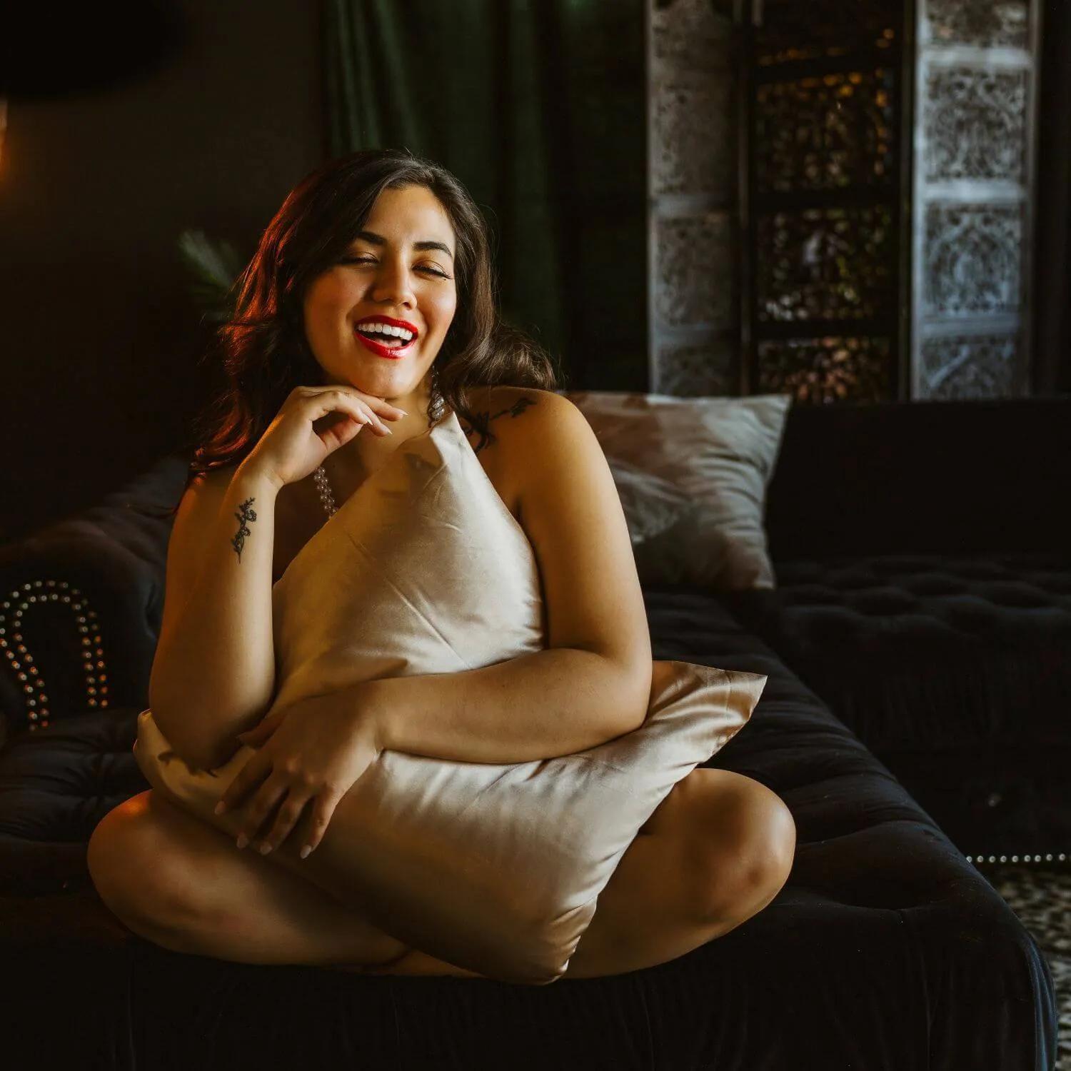 Red-lipped woman sitting crisscross with pillow in lap laughing.