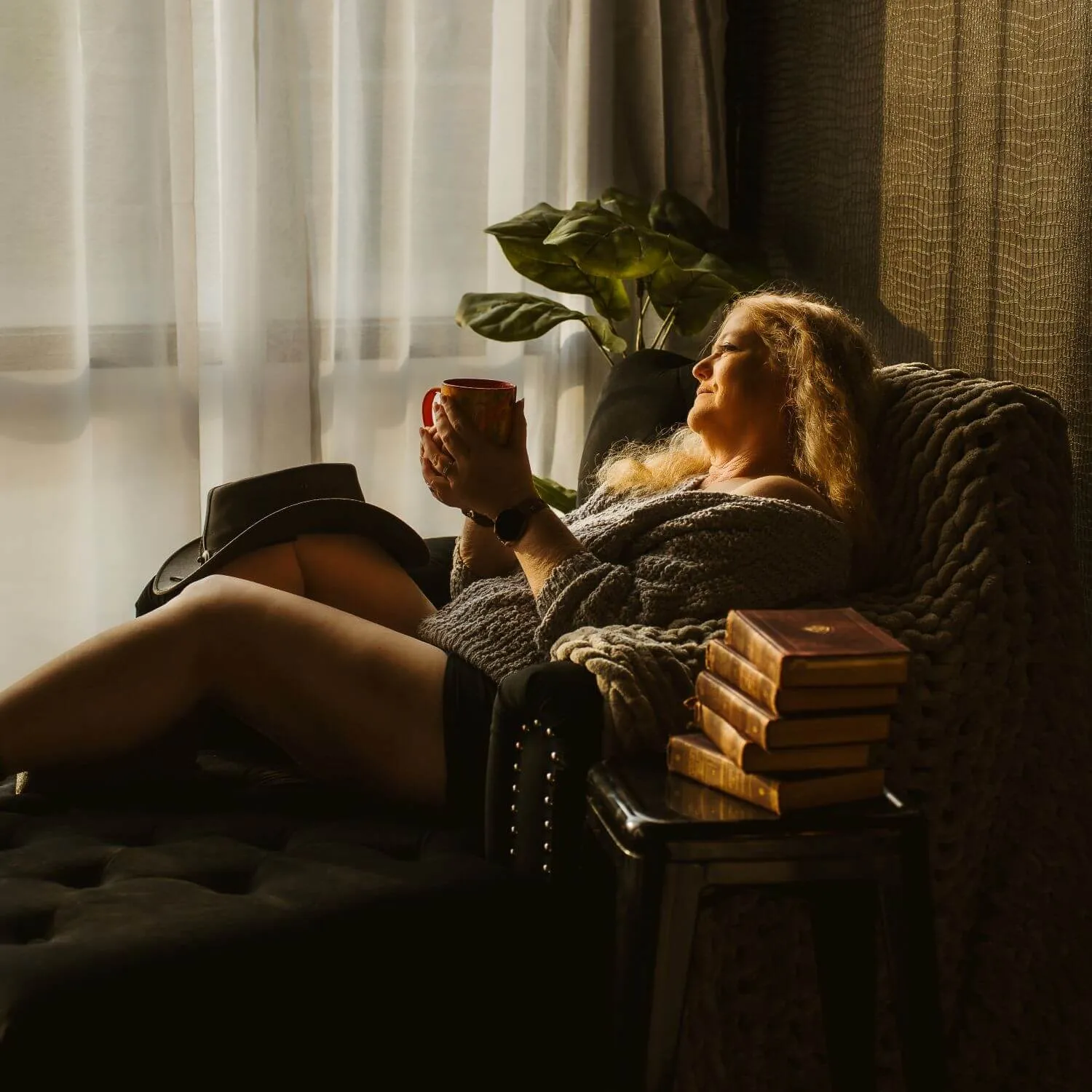 Senior blonde, lounging, holding coffee in hands as she gazes out of the window; cowgirl hat resting on knee. 