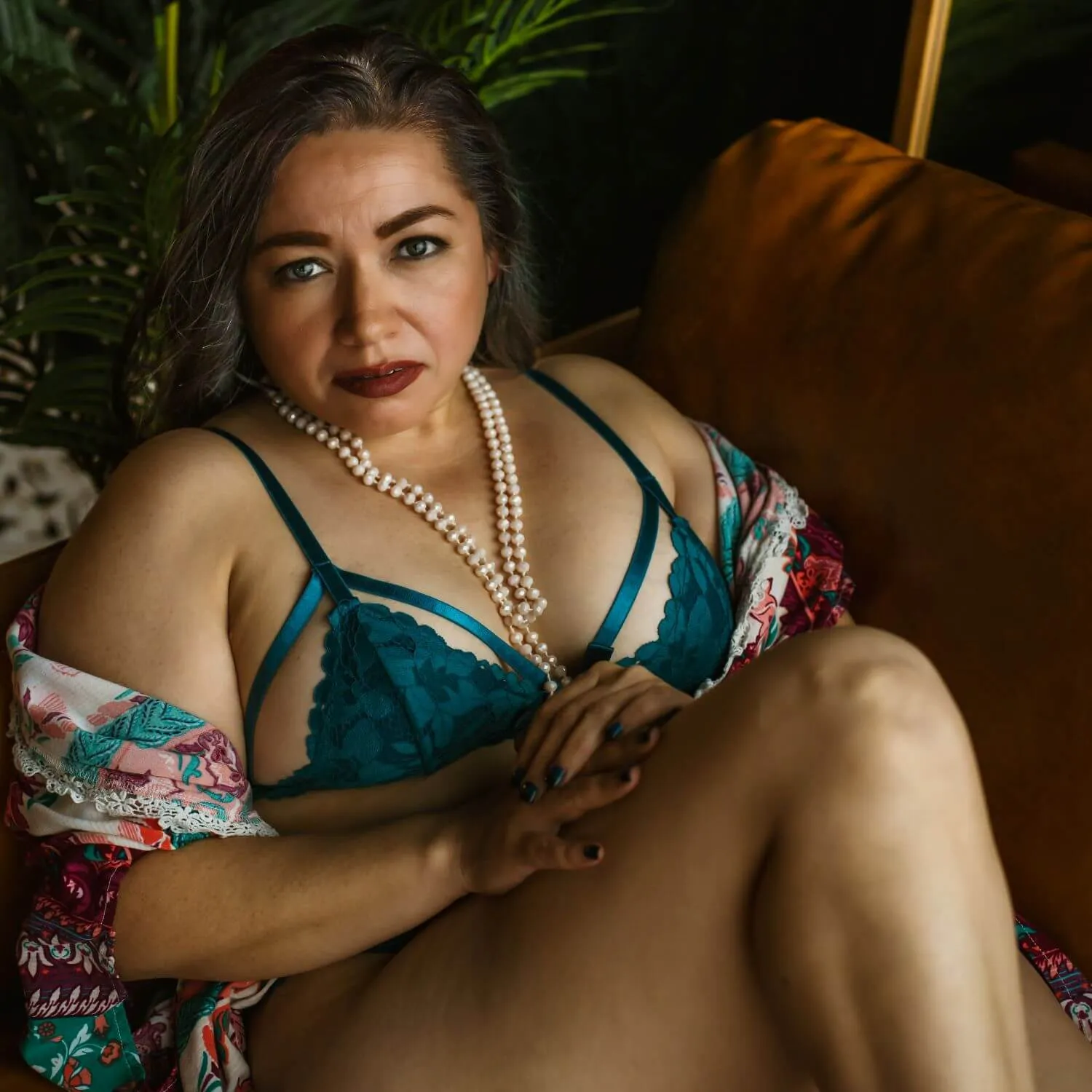 Mature woman gazing confidently at camera in teal lingerie and pearls.