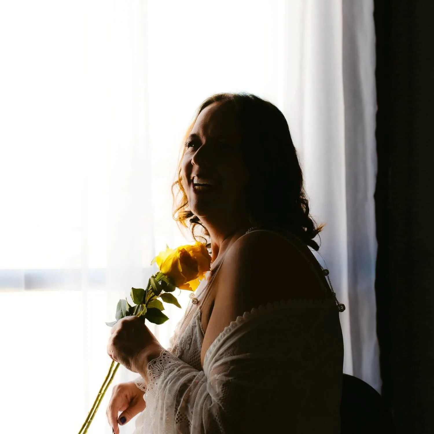 Woman laughing in white lace, holding yellow flower close to chest. 