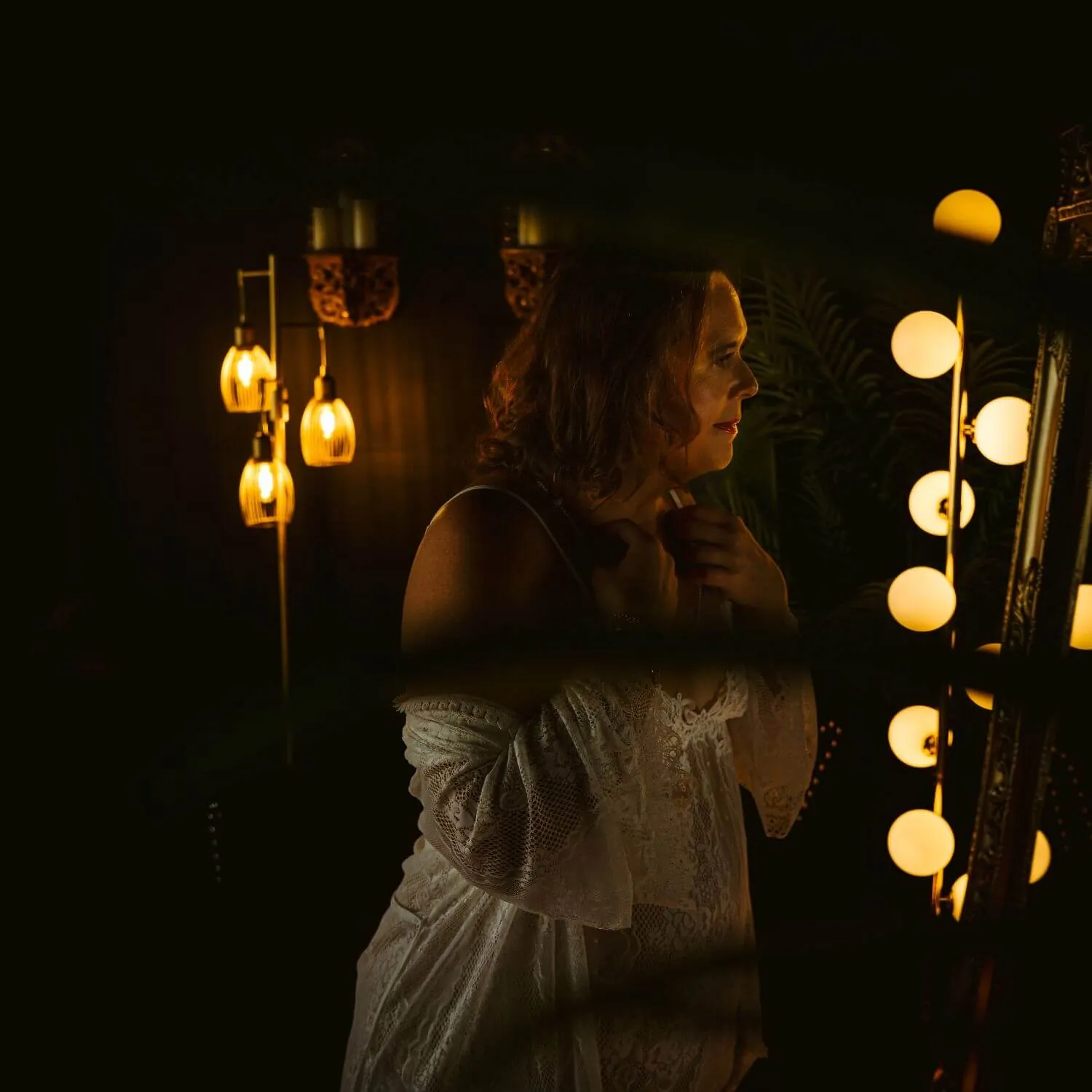 Woman in white lace, adjusting jewelry as she smiles into mirror.