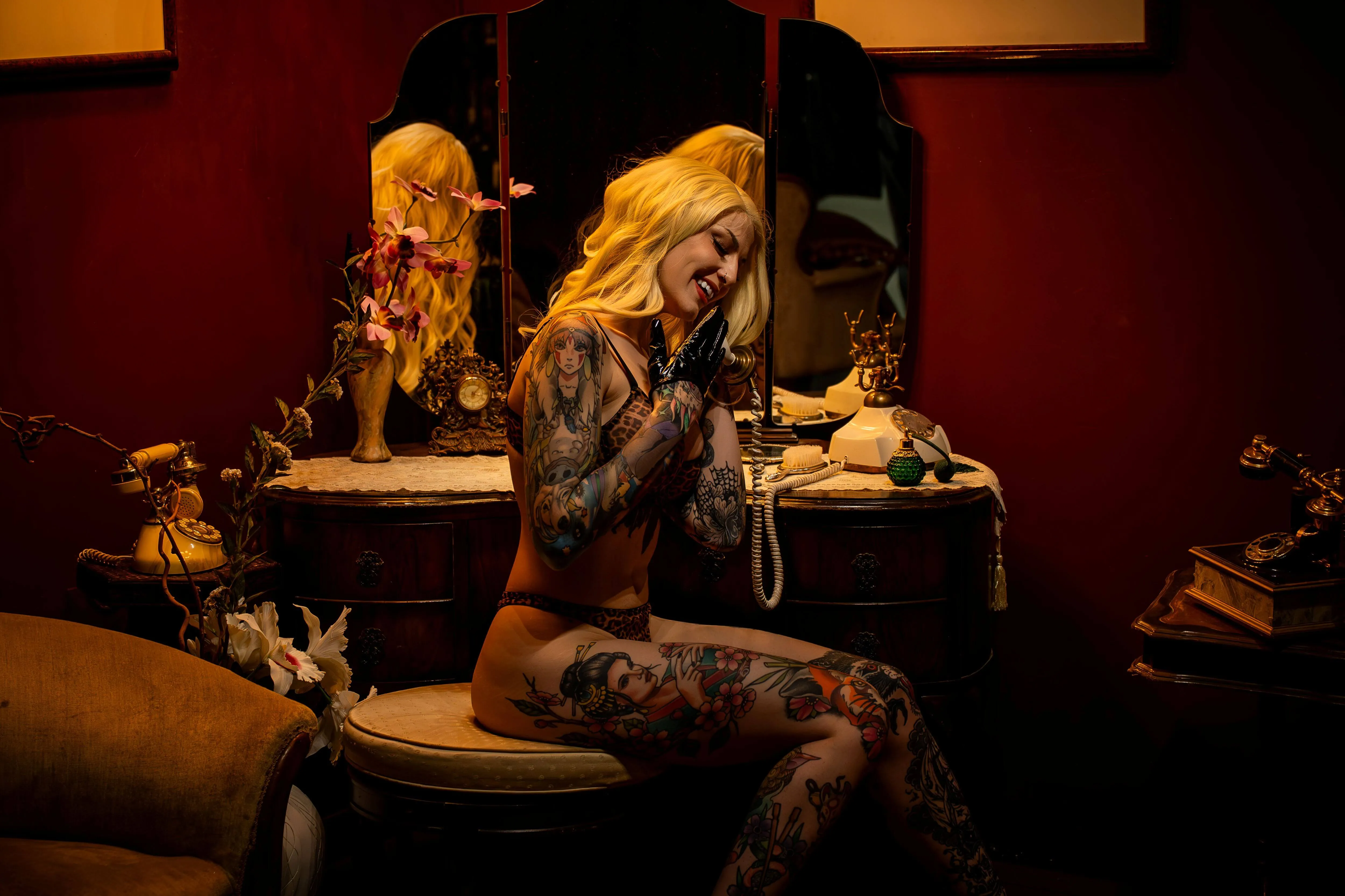 Young tattooed blonde woman in black lingerie, smiling while sitting at vintage boudoir vanity; talking on vintage phone.