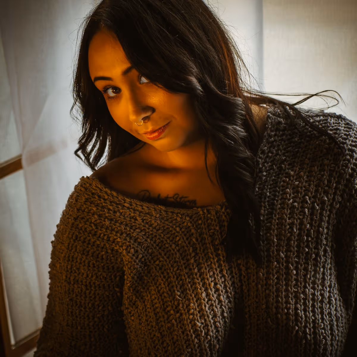 Woman with long dark hair wearing a brown knitted sweater and a septum nose ring, looking confidently with soft lighting.