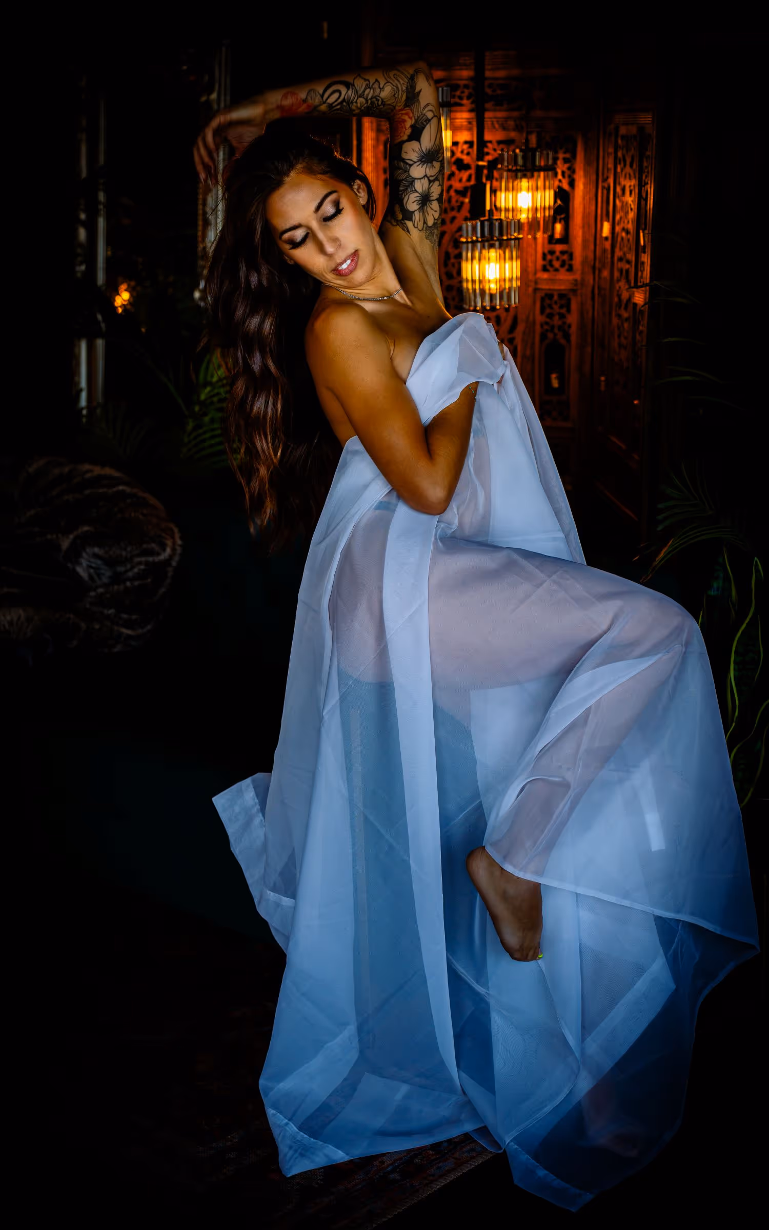 Woman with long hair and floral arm tattoo posing wrapped in a sheer white fabric in dimly lit room with warm lights in the background.