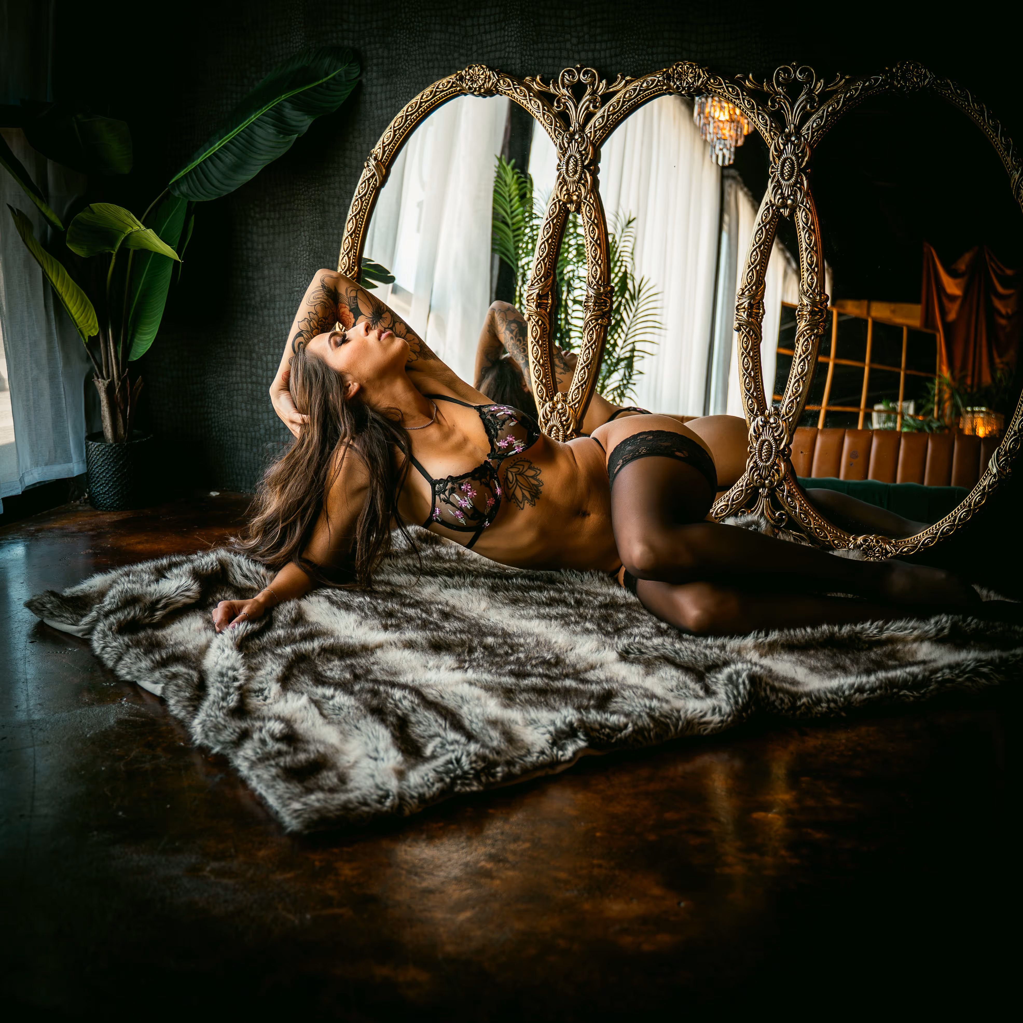 Woman in black lingerie with tattoos posing on a gray faux fur rug in front of a large ornate gold-framed mirror indoors.