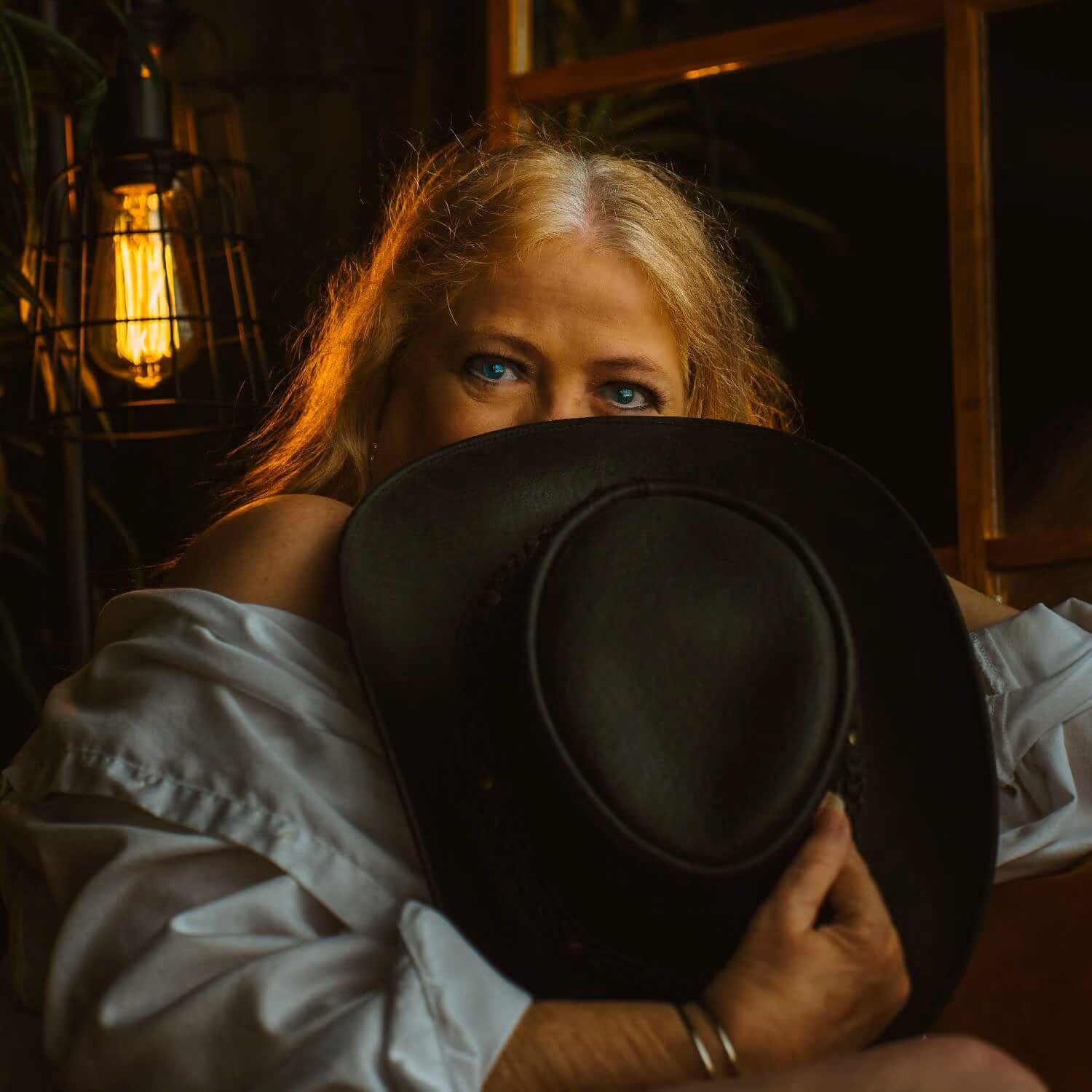 Senior boudoir set, in unbuttoned light blue shirt, playfully hiding face with black cowgirl hat.