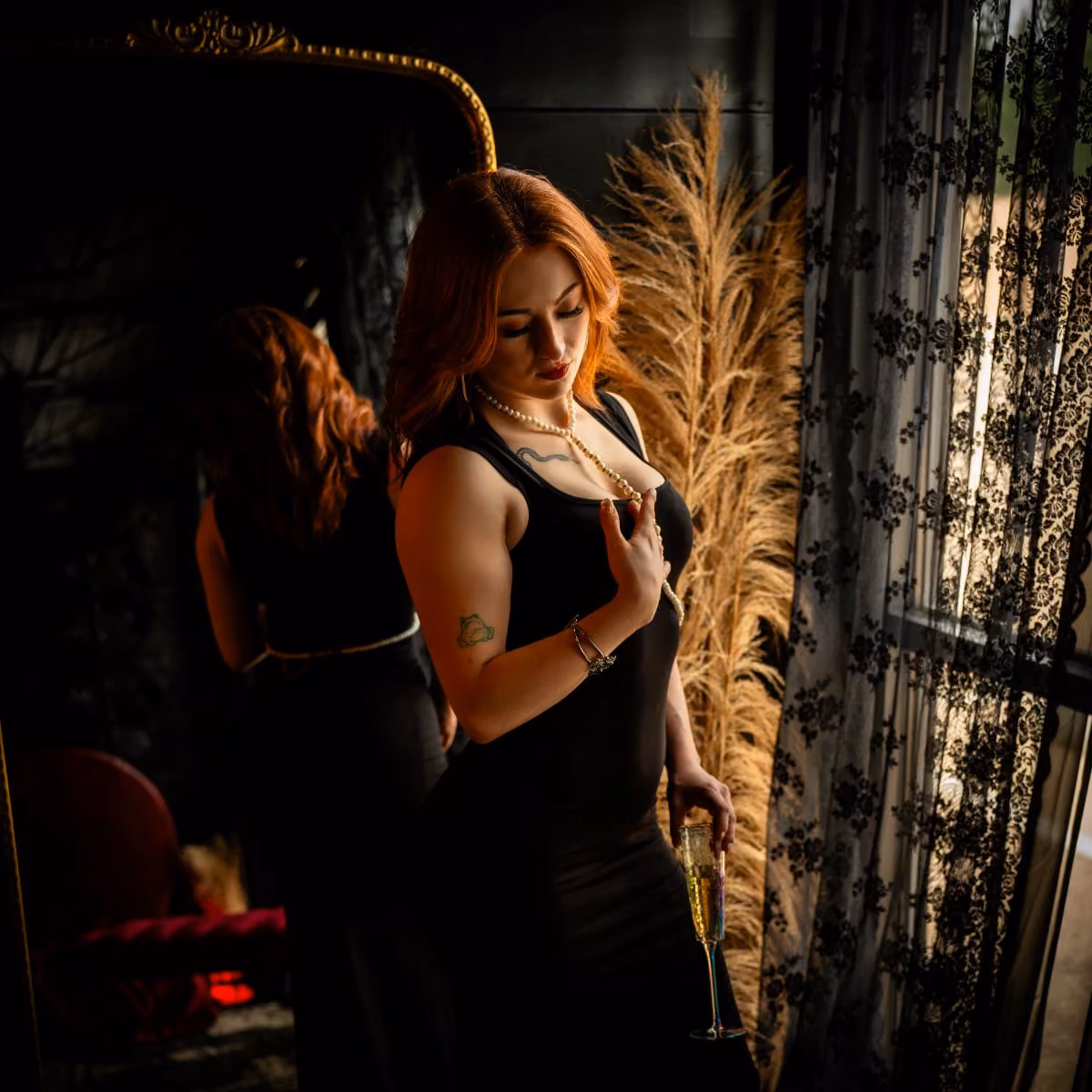 Woman in a black dress holding a champagne flute and a pearl necklace, by a window.