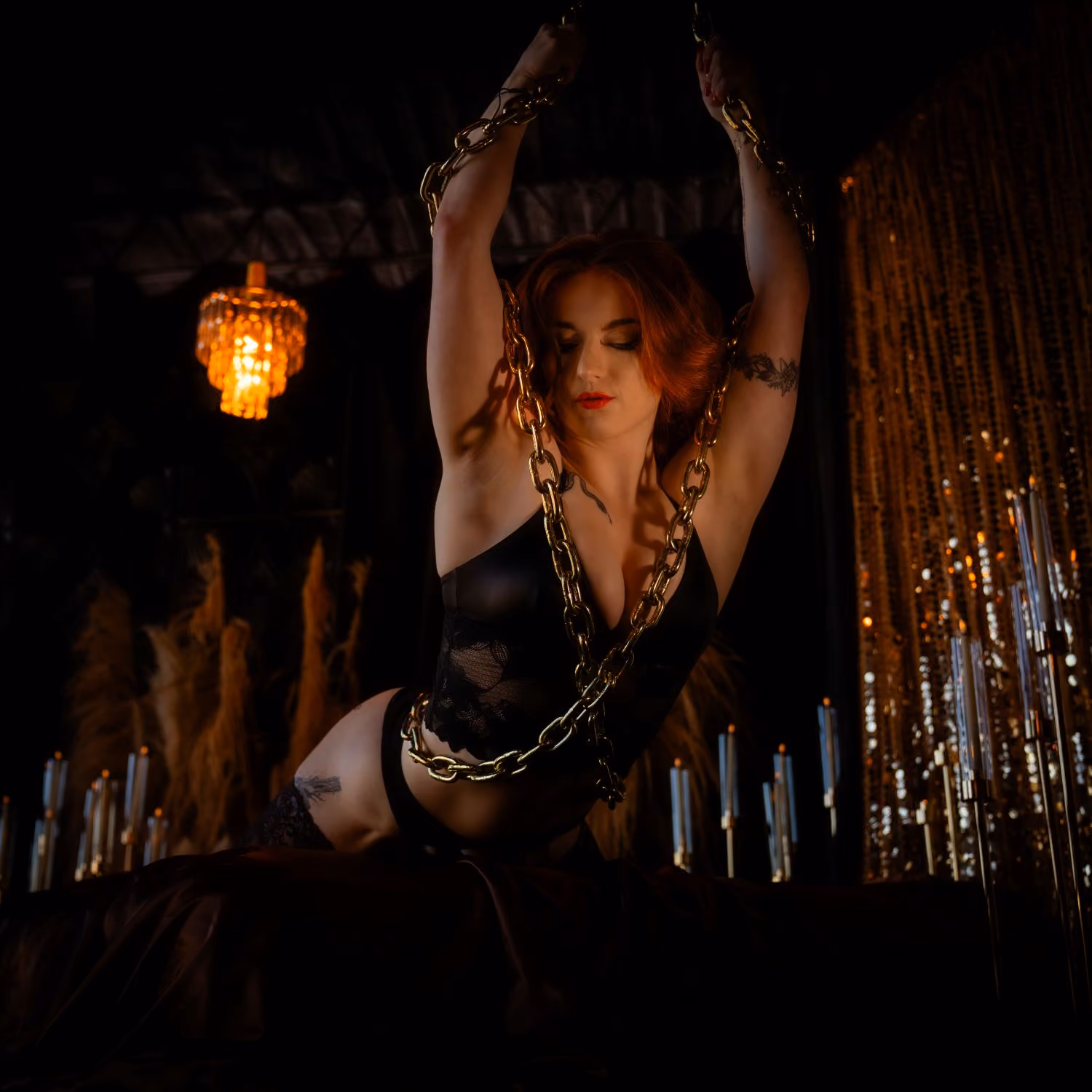 Red-haired woman in black lingerie posing with gold chains, eyes closed, candlelit room.