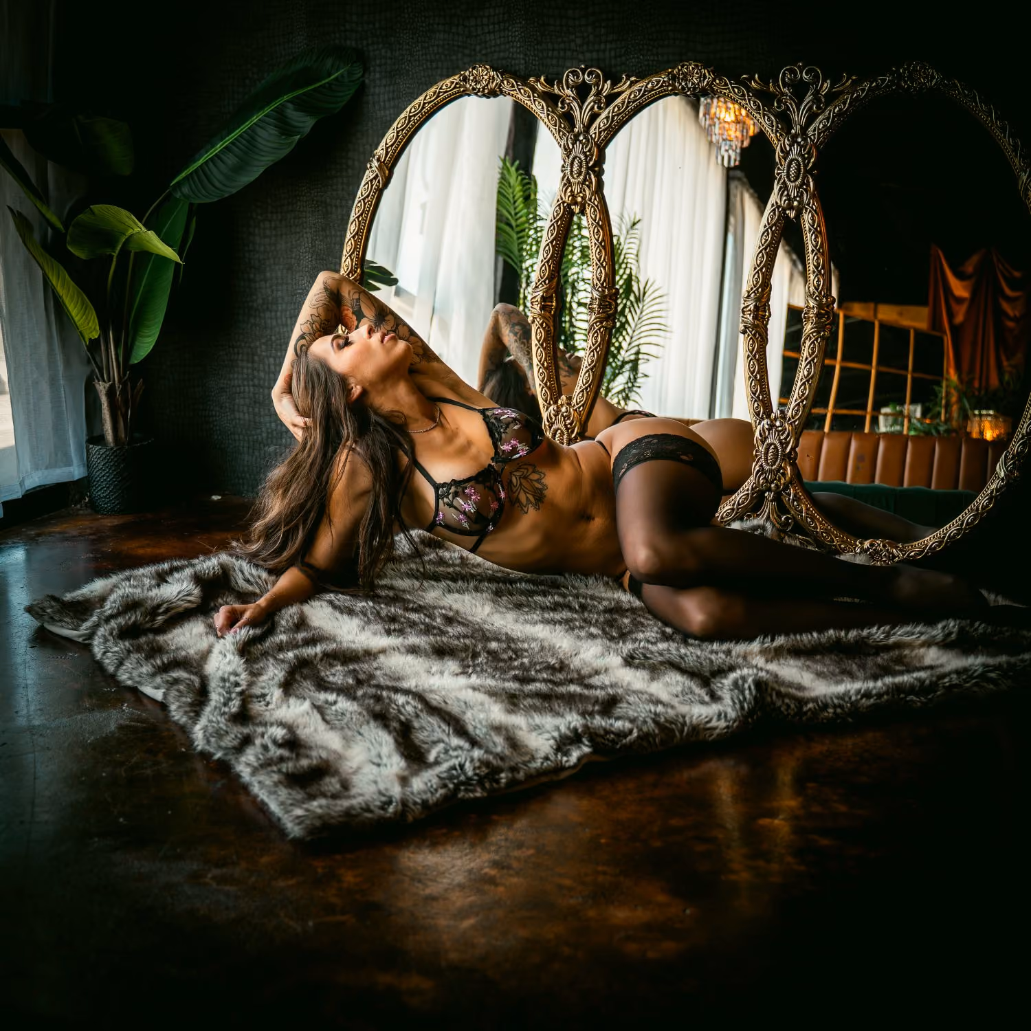 Tattooed woman reclining on a faux fur rug before three ornate mirrors, dark interior.