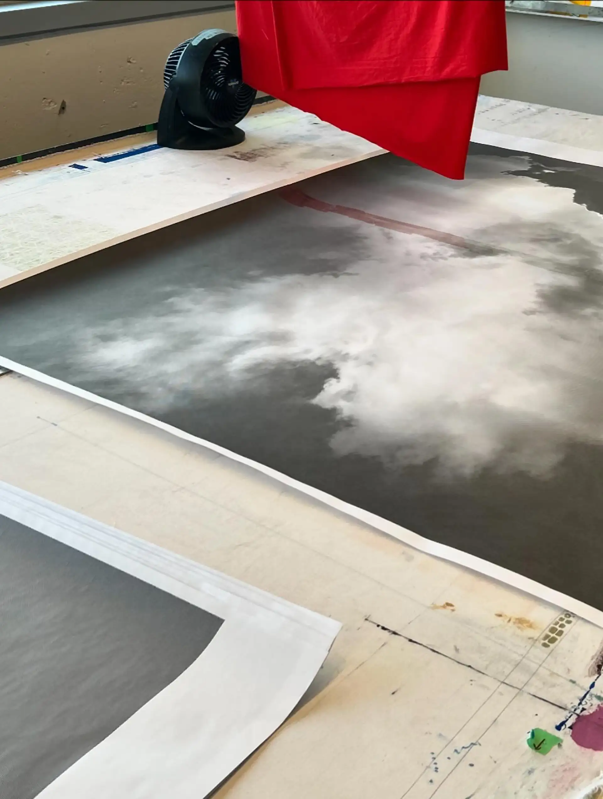 A large-format monochrome cloud print lies on a flat surface, partially covered by a red cloth suspended above it, with a fan nearby aiding the drying process. Impression grand format en noir et blanc représentant des nuages, posée sur une surface plane, partiellement couverte par un tissu rouge suspendu, avec un ventilateur à proximité pour faciliter le séchage.