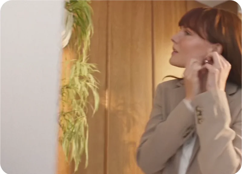 A woman adjusts her earring while getting ready to leave, framed in warm morning light against a wooden wall—capturing a moment of quiet preparation. / Une femme ajuste sa boucle d’oreille avant de partir, baignée dans une lumière matinale chaude, devant un mur en bois — un instant de préparation calme.