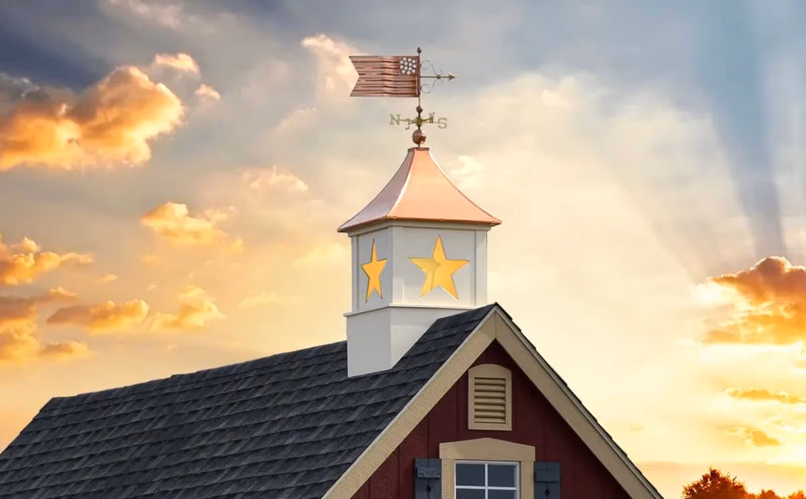 Designer Series star-cutout cupola with glowing yellow light and copper roof mounted on a red barn at sunset