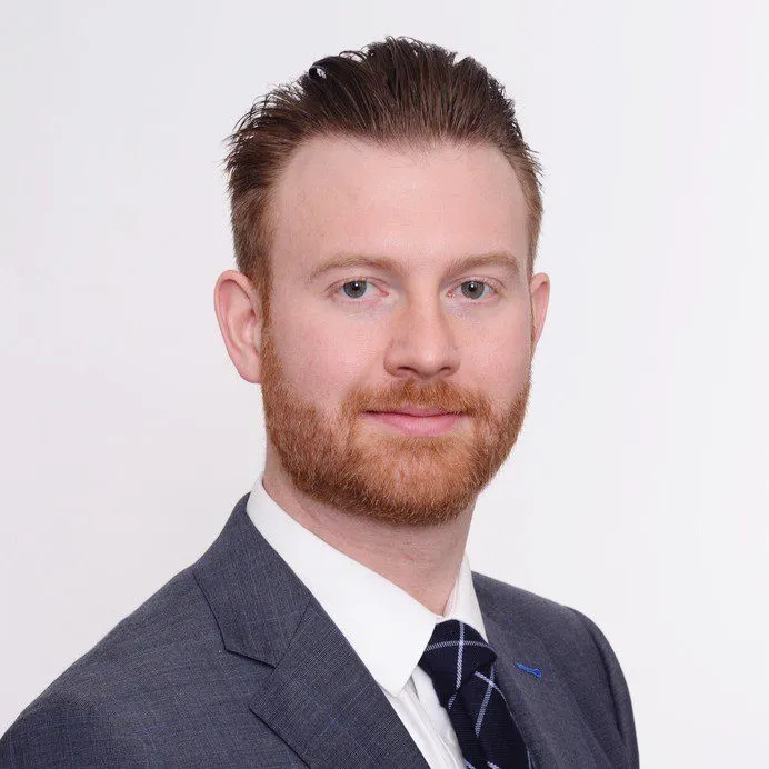 Portrait of a man with red hair and beard wearing a gray suit and striped tie against a white background.
