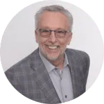Smiling older man with gray hair and glasses wearing a gray checkered blazer and light blue shirt against a plain background.