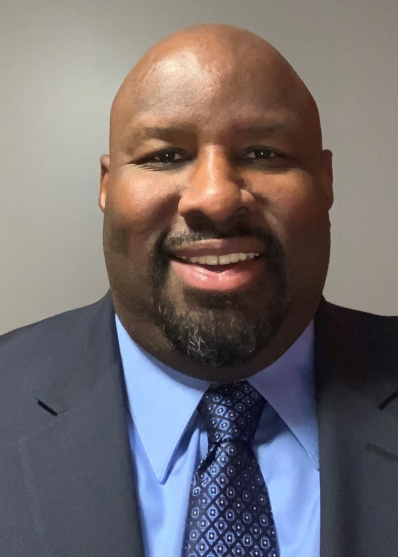 Smiling bald man with a goatee wearing a dark suit, light blue shirt, and patterned blue tie against a plain background.