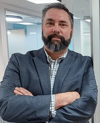 Bearded man with dark hair wearing a dark blazer and checkered shirt standing with arms crossed in a modern office.