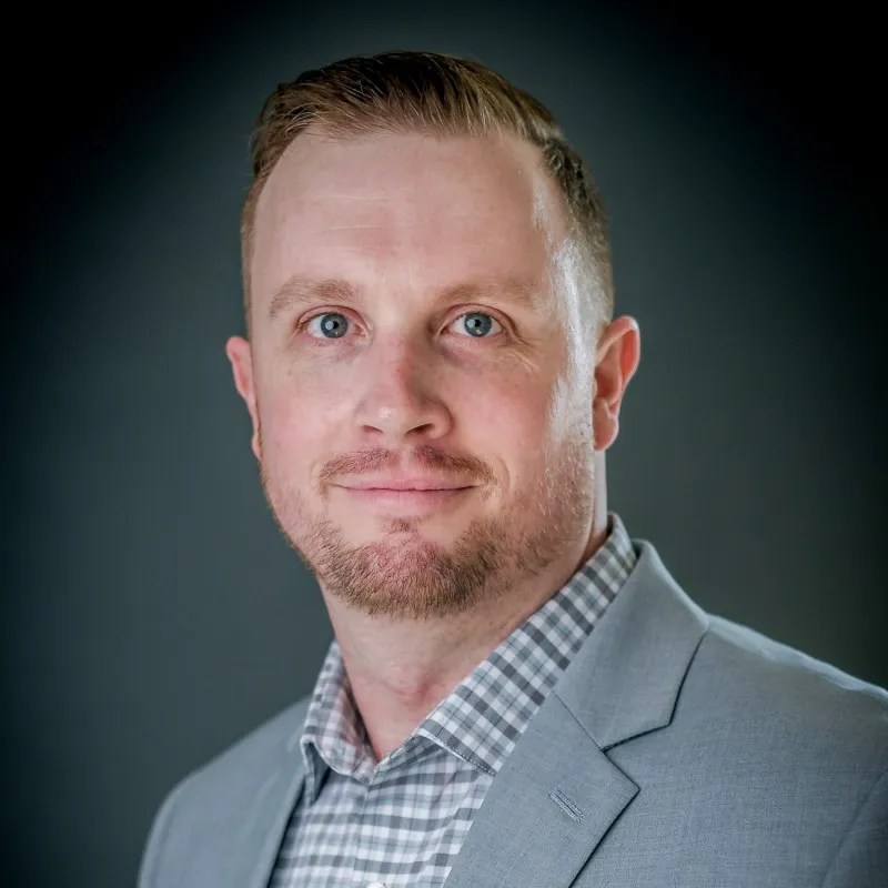 Portrait of a man with light skin and short hair wearing a gray blazer and checkered shirt against a dark blurred background.