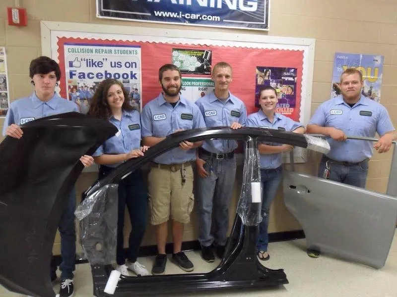 Five collision repair students in blue uniforms standing indoors holding different car parts including a black car door frame and a silver car door.