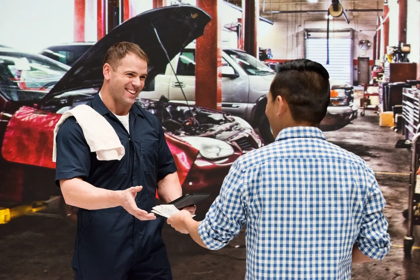 Smiling mechanic in a dark blue uniform with a towel over his shoulder receiving cash payment from a customer in a blue checkered shirt inside an auto repair shop.