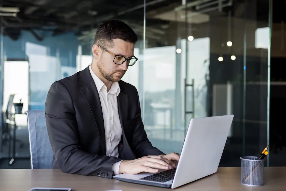 Man in business attire typing on a laptop at a wooden desk in a modern office with glass walls.