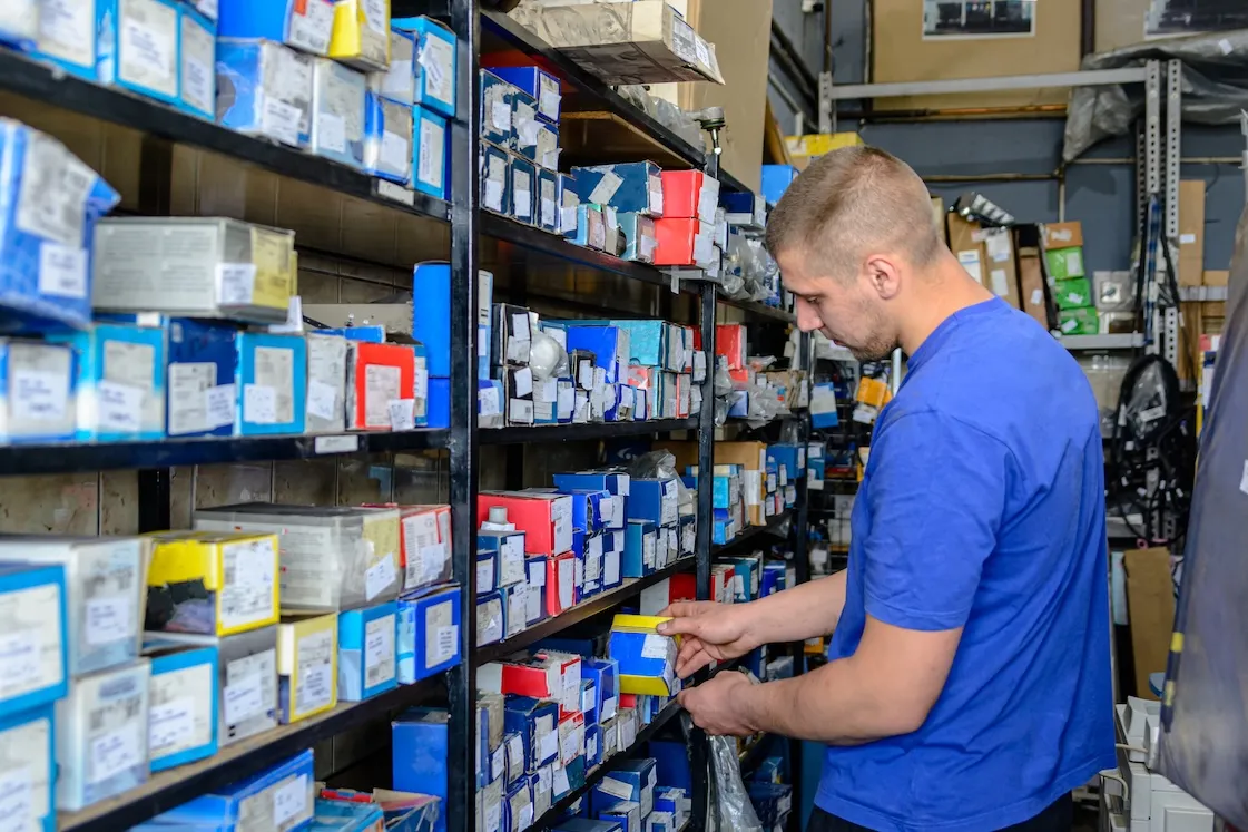 Man in a blue shirt selecting a small boxed item from metal shelves filled with various auto parts in a storage room.