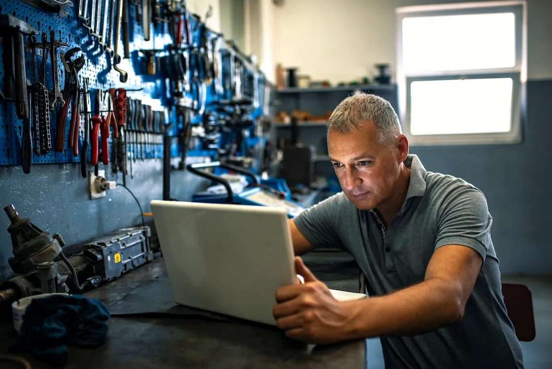 Man in gray polo shirt working on a laptop in a workshop with tools hanging on a pegboard wall.