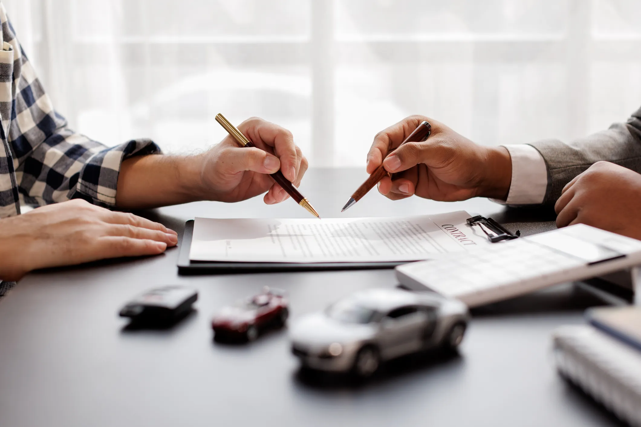Two people holding pens over a contract document on a table with toy cars and a calculator nearby.