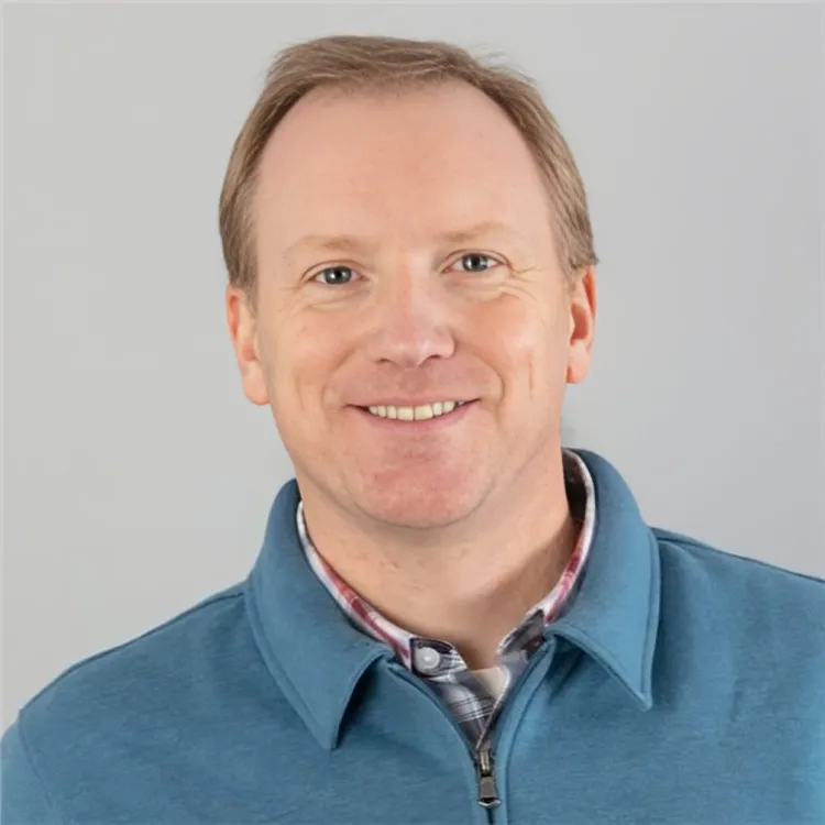 Smiling middle-aged man with light brown hair wearing a blue zip-up sweater over a plaid shirt against a plain light gray background.