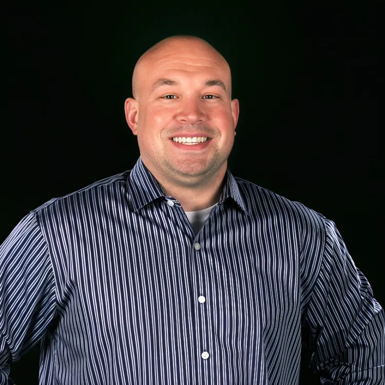 Smiling bald man with hands on hips wearing a striped button-up shirt against a black background.