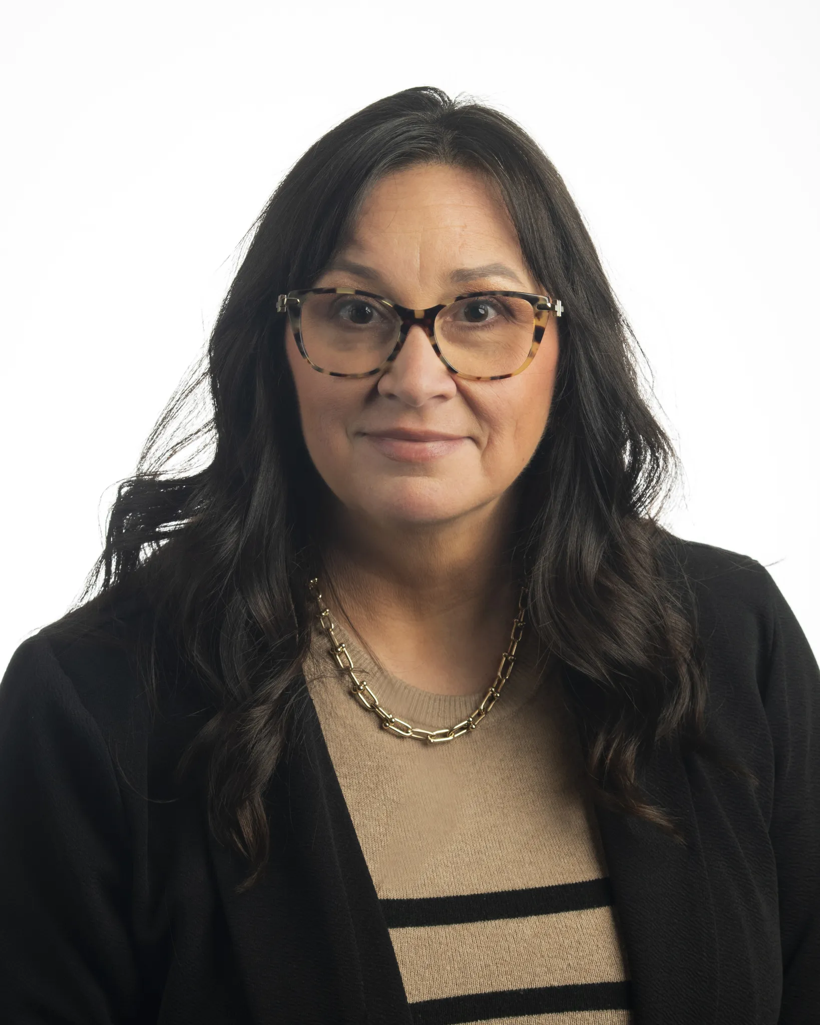 Woman with long dark hair wearing tortoiseshell glasses, a gold chain necklace, a beige sweater with black stripes, and a black blazer, against a white background.