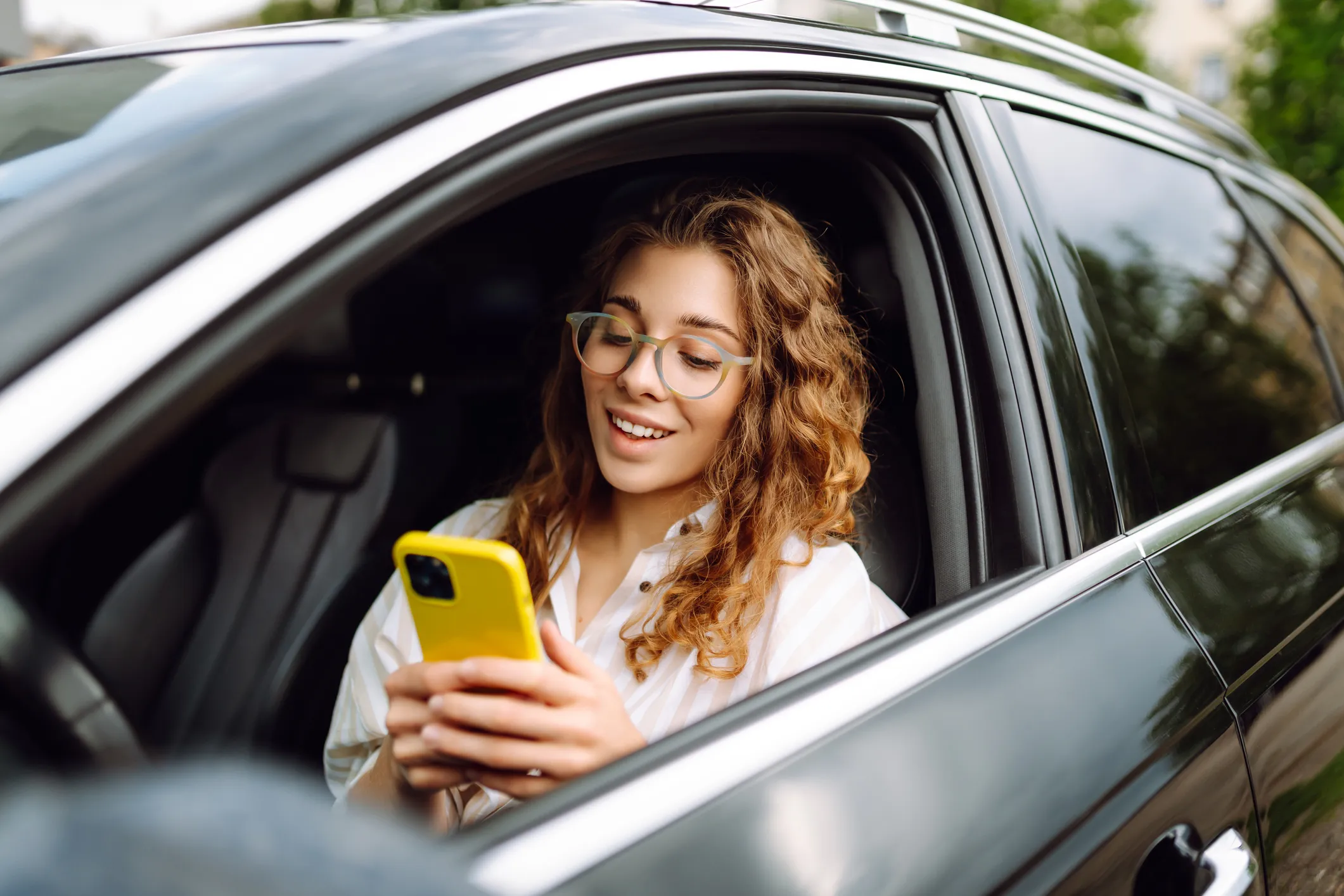 Smiling woman with curly hair and glasses sitting in a black car using a yellow smartphone.