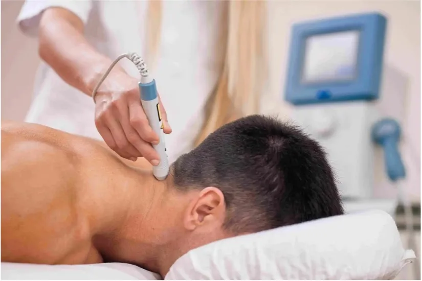 Person receiving a neck treatment with a handheld medical device while lying face down on a white pillow.