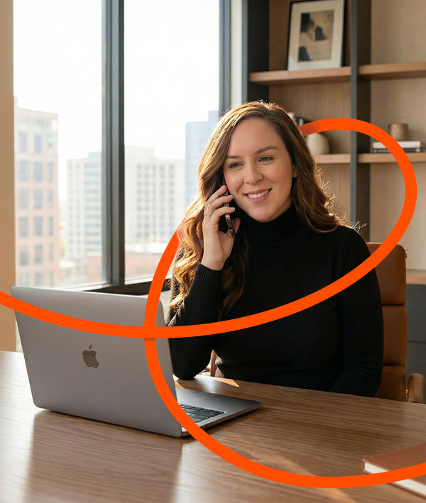 Smiling woman in black turtleneck sitting at a desk with a laptop, talking on a phone in a bright office.