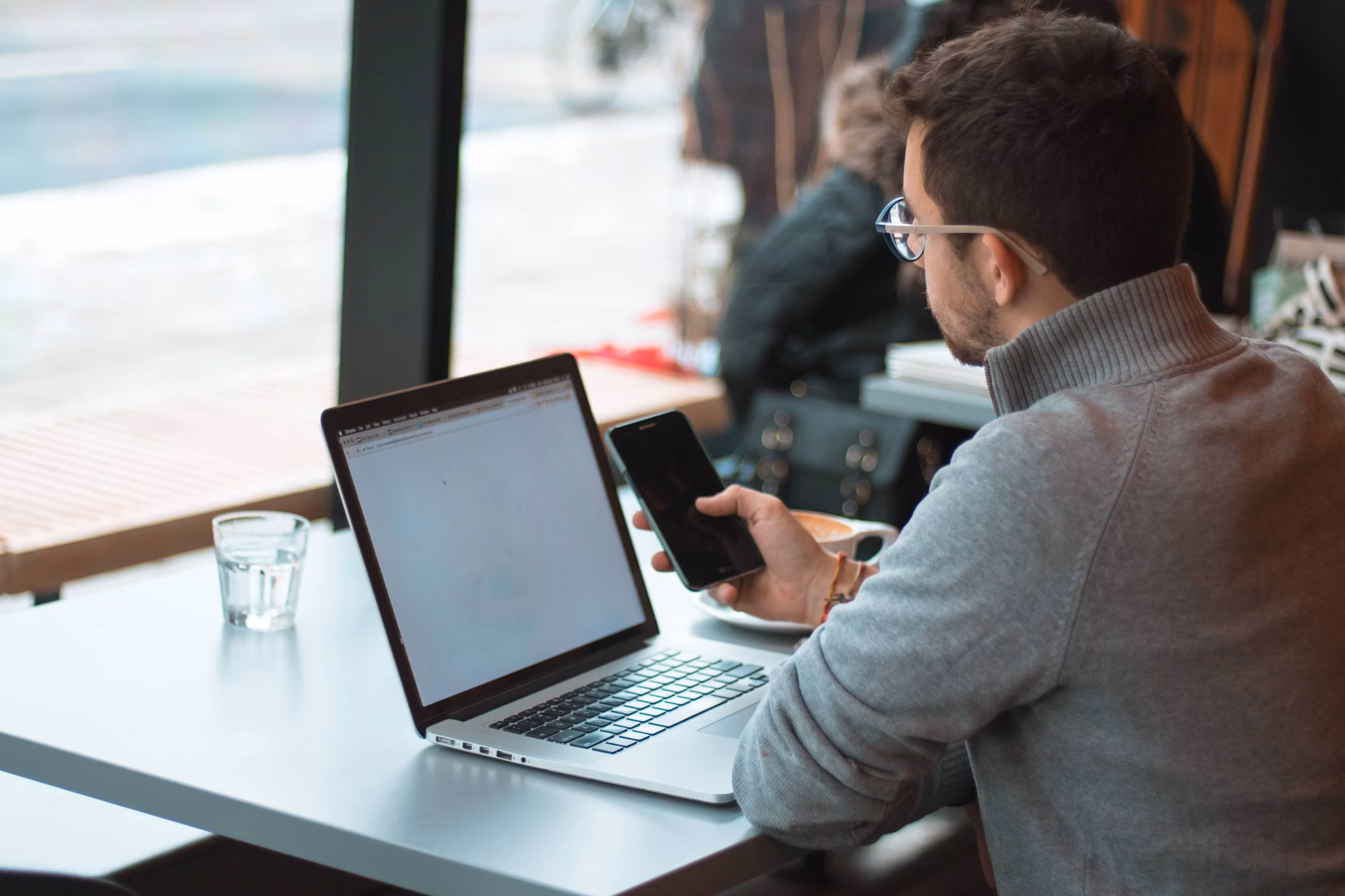 Man sitting at a café table working on a laptop while holding a smartphone, with a coffee cup nearby.