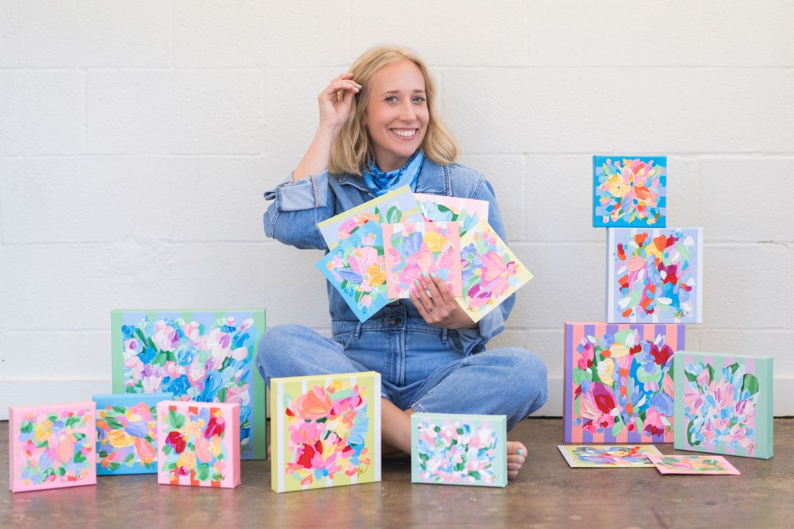 dfw artist jessa adler of jessa adler art sits between her pastel and floral art pieces while holding several small paintings in front of white wall
