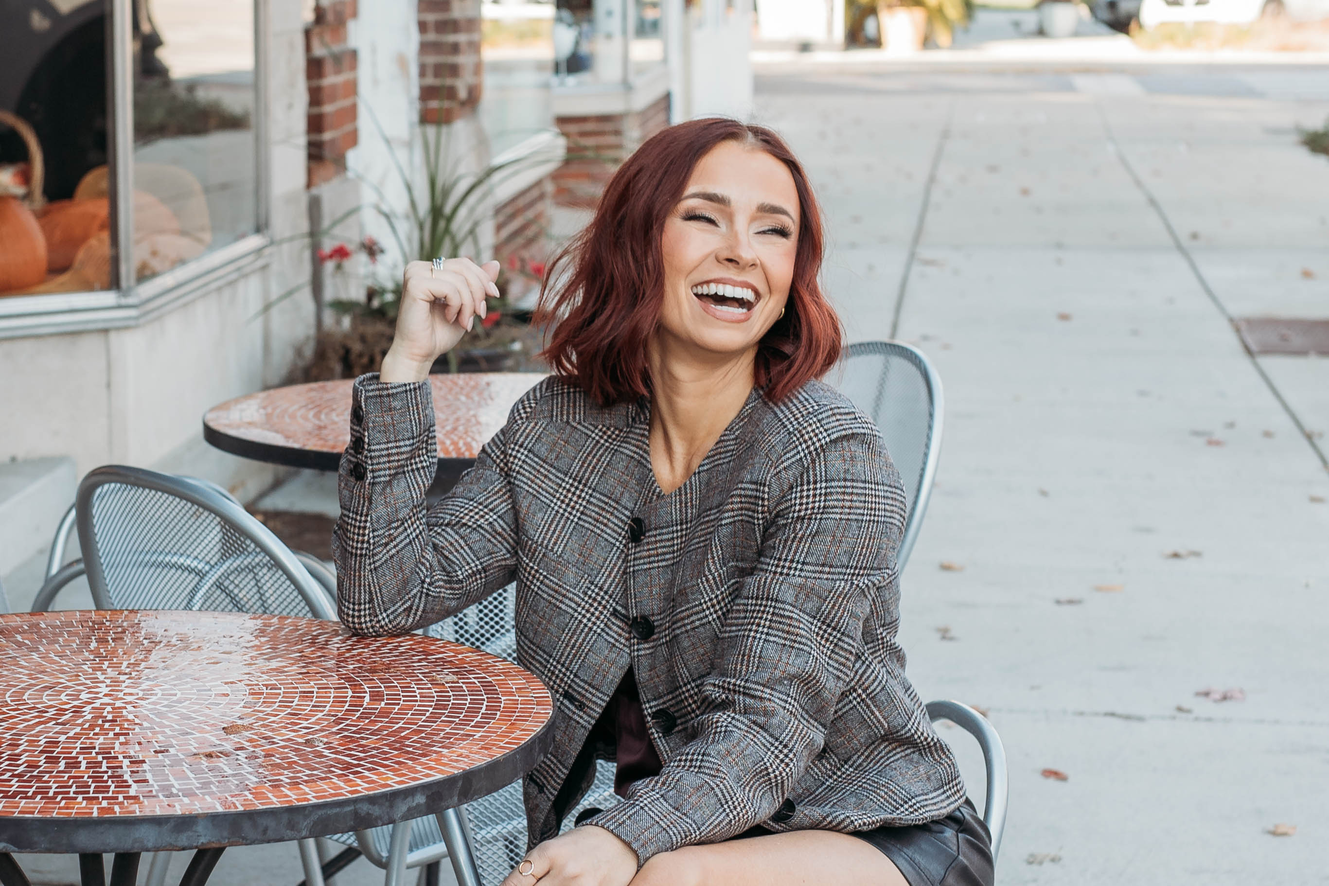 Smiling woman with short red hair sitting at an outdoor café table, wearing a plaid jacket and laughing on a city sidewalk