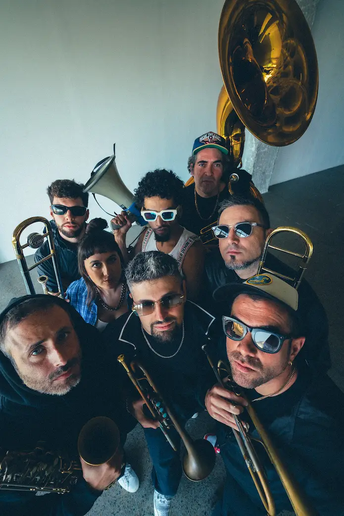Eight musicians wearing sunglasses pose with brass instruments including trumpets, trombones, a tuba, and a saxophone in an indoor setting.