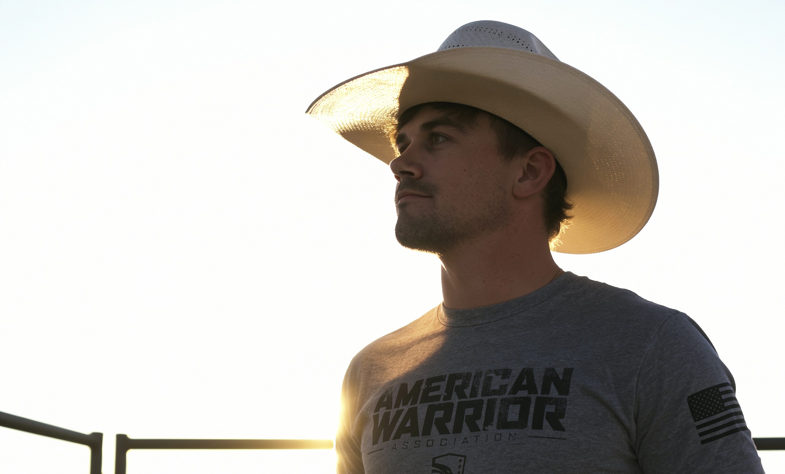 A bull rider stands in an open field at dusk wearing an American Warrior Association shirt.