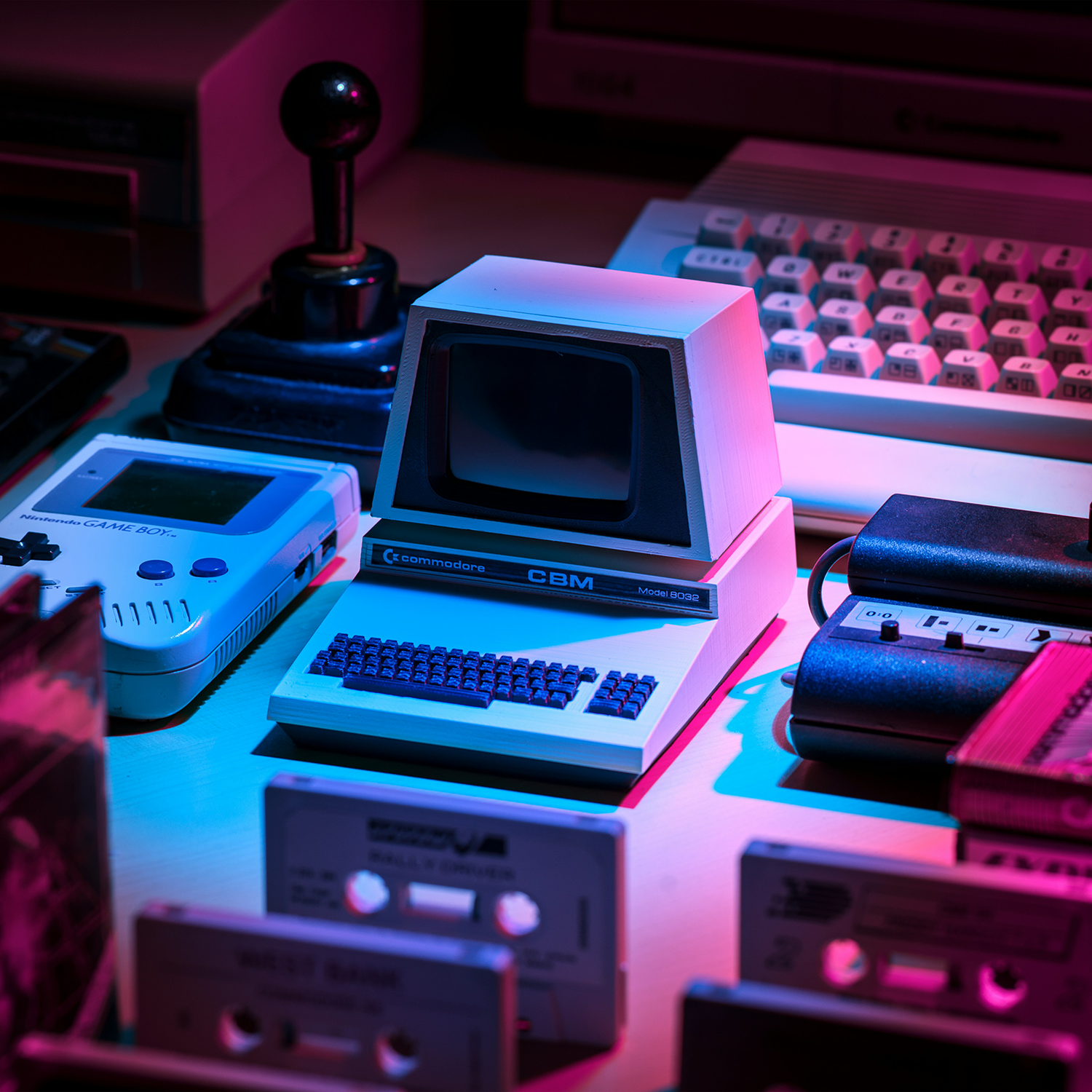 A vintage Commodore CBM Model 8032 computer surrounded by cassette tapes, a Nintendo Game Boy, and retro gaming accessories under purple and blue lighting.