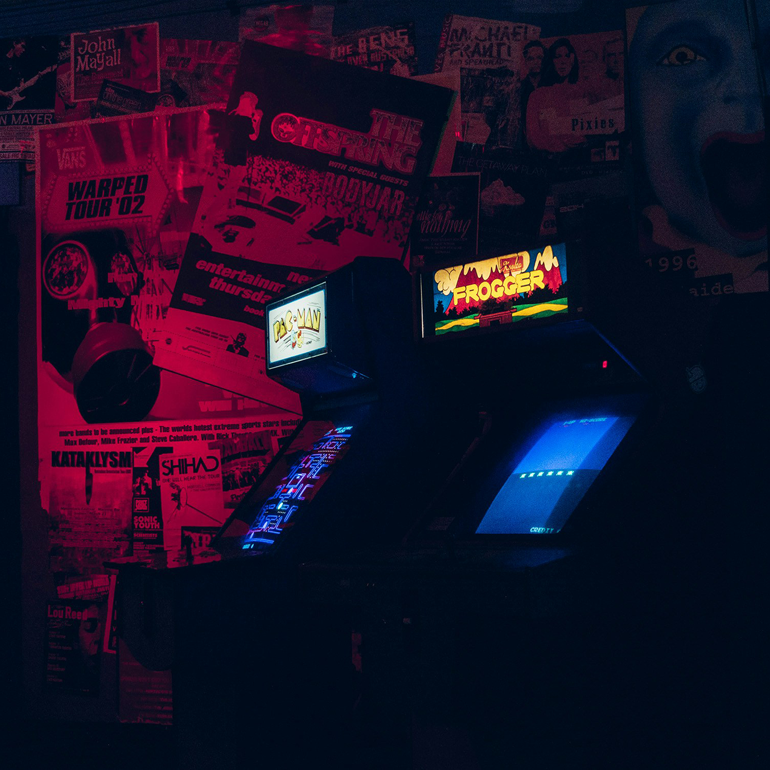 Dimly lit room with vintage arcade machines displaying Pac-Man and Frogger games, red-lit wall covered with posters in the background.