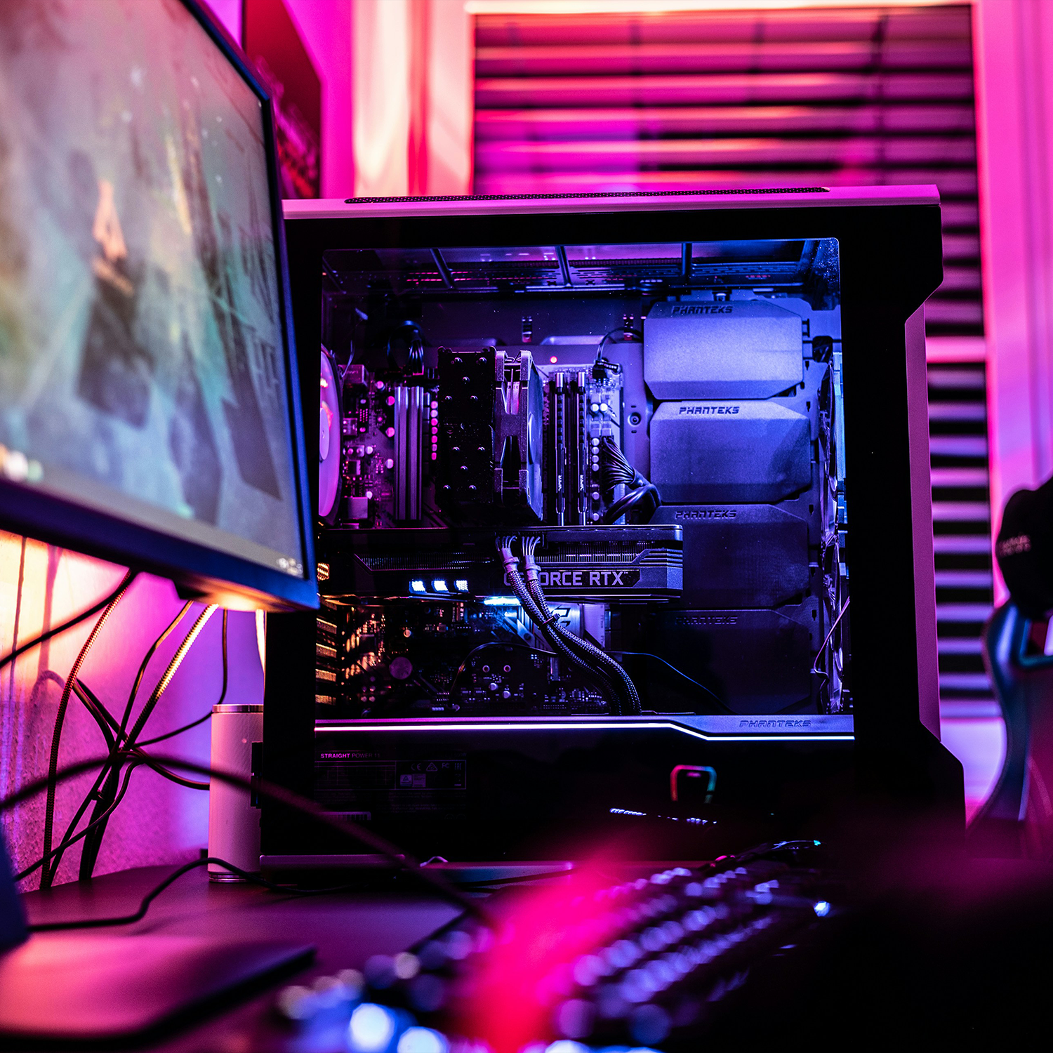 Gaming PC setup with monitor displaying a game and a transparent PC case illuminated with purple and blue LED lights showing internal components including a GeForce RTX graphics card.