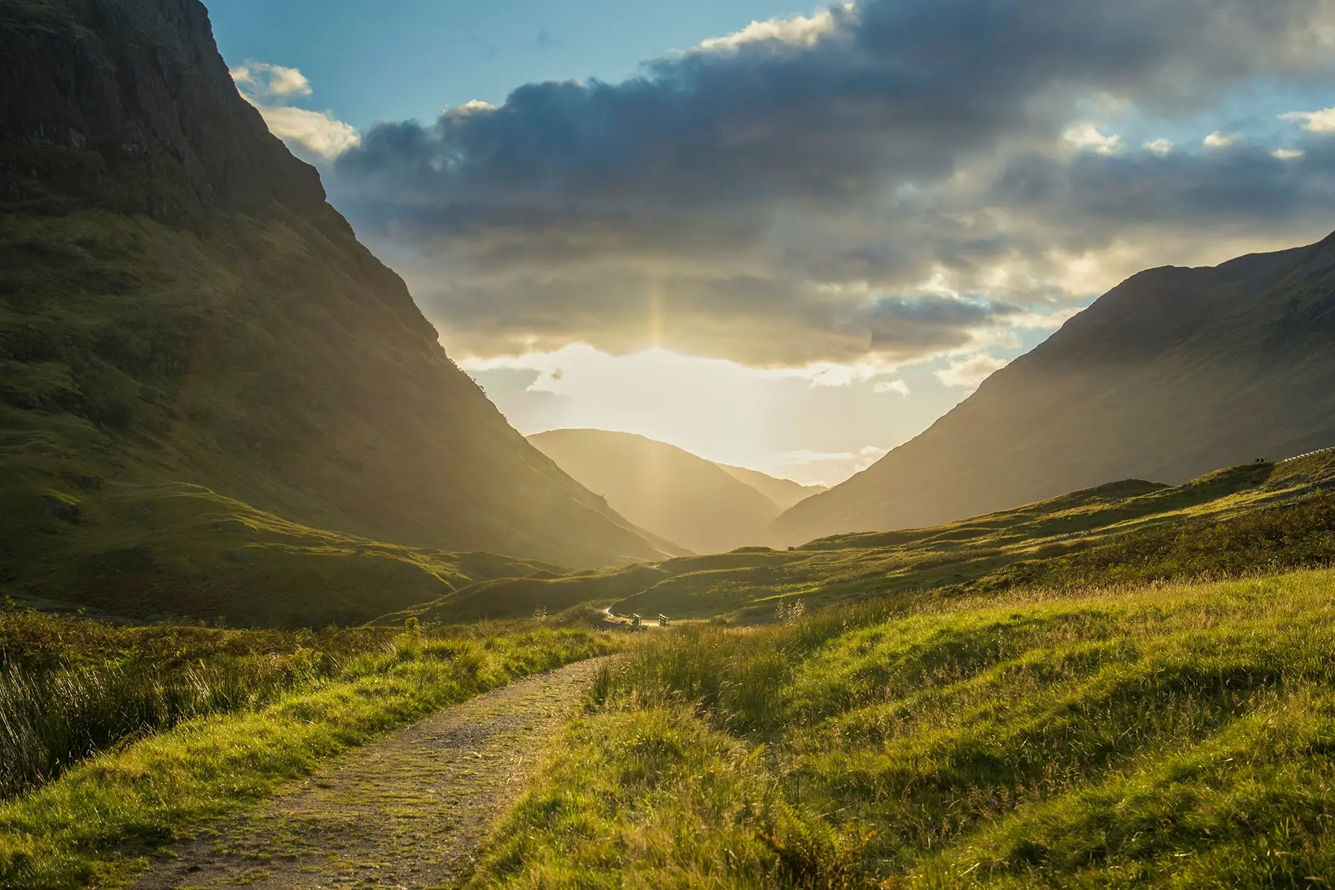Sunset over a grassy valley with a winding path between tall hills under partly cloudy sky.