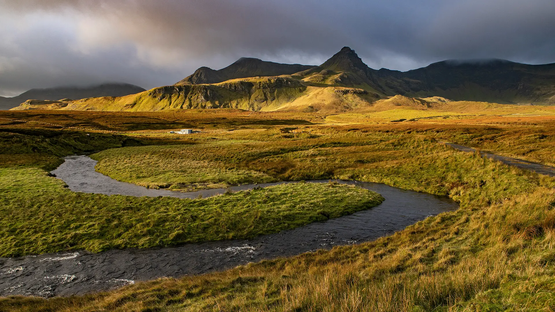 Winding river through grassy fields with rugged mountains and moody clouds in the background.